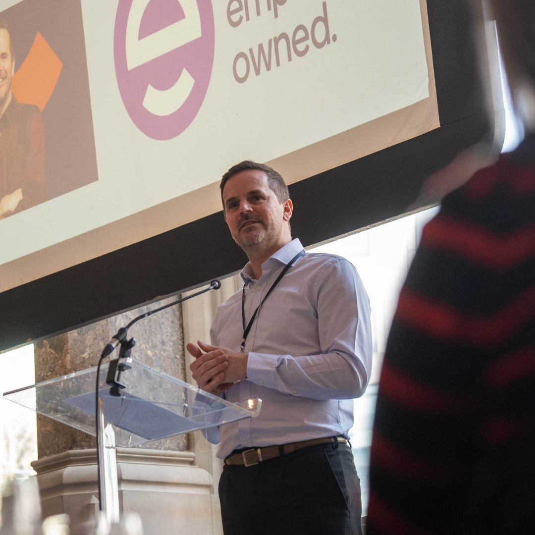 James de la Vingne, Employee Ownership Association James de la Vingne from the Employee Ownership Association speaking at the Aspire Banking Hall in front of a projector screen.
